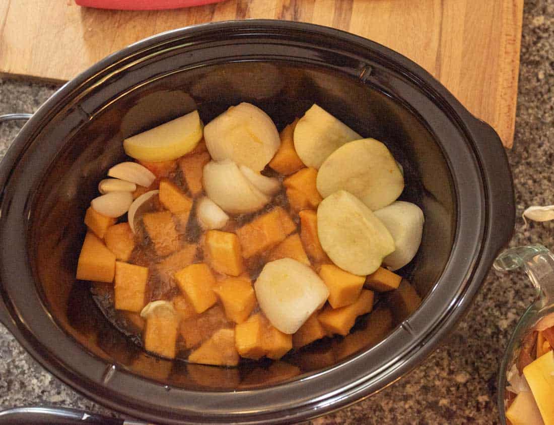 A slow cooker filled with cubed butternut squash, chunks of onion, sliced apples, and whole garlic cloves, ready to be cooked.
