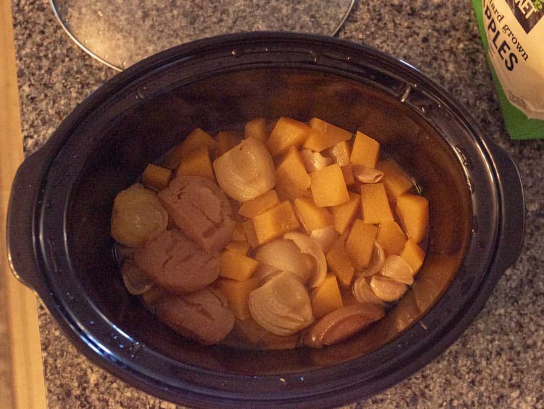 A slow cooker on a countertop filled with chunks of yellow squash, halved onions, and several brown sausages being cooked together.