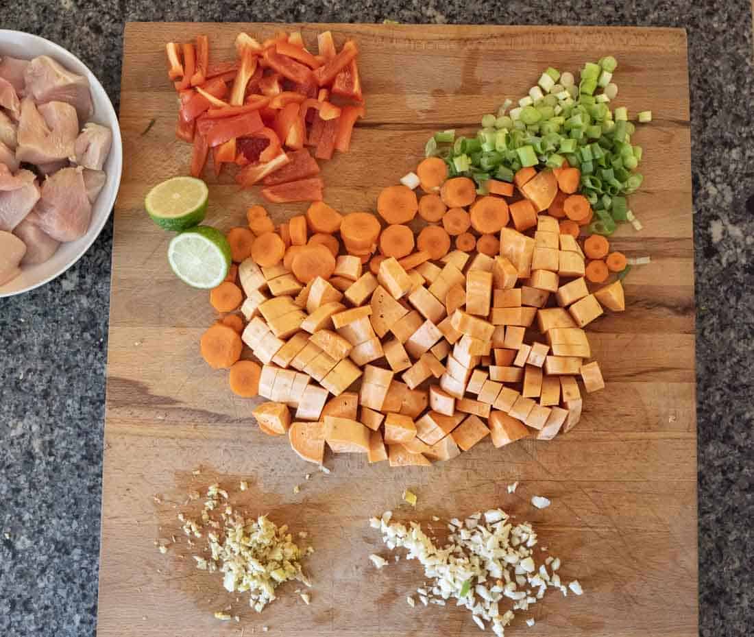Chopped sweet potatoes, carrots, green onions, and red bell peppers are arranged on a wooden cutting board, with minced garlic and ginger in small piles and a halved lime. A plate of raw chicken sits to the side.