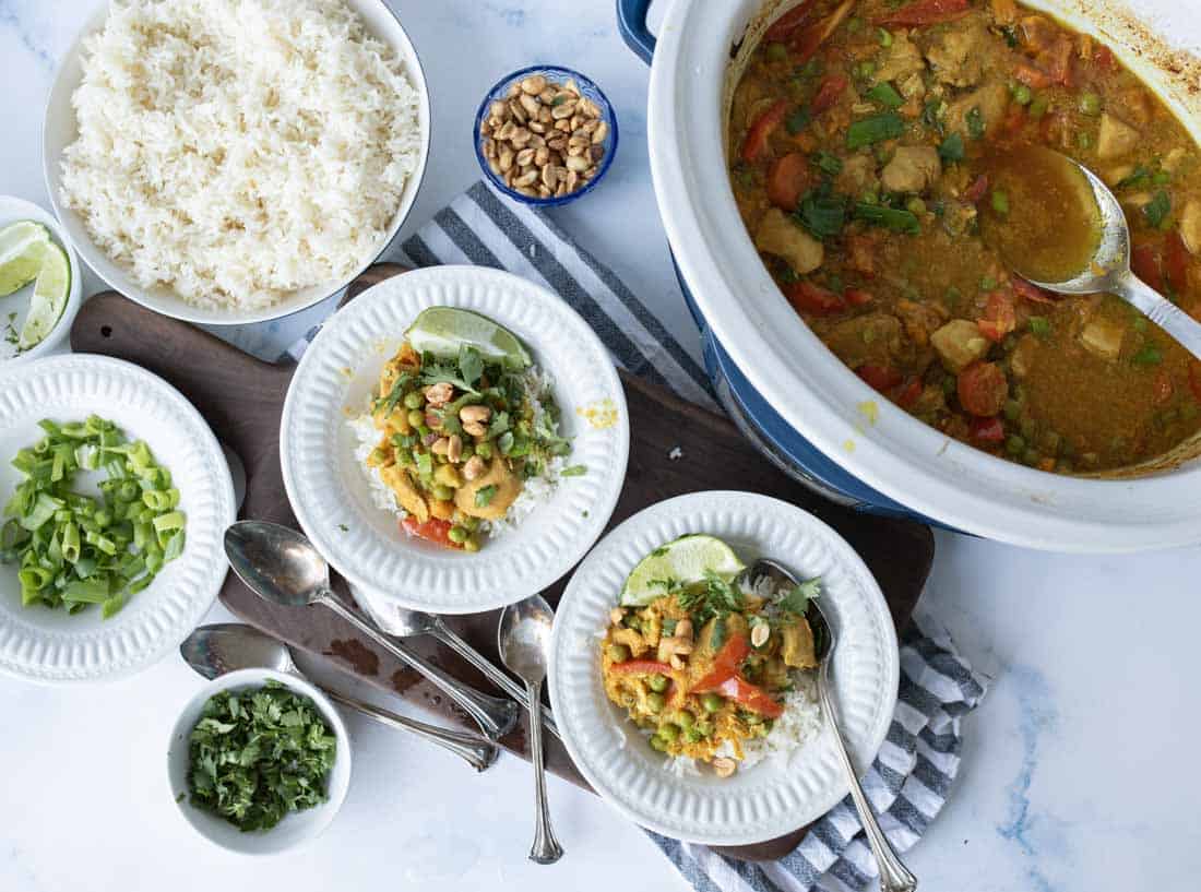A table set with bowls of rice, a curry dish in a pot, plates of curry over rice, and small bowls of chopped green onions, cilantro, peanuts, and lime wedges, plus several serving spoons.