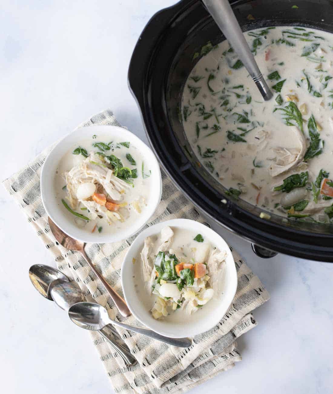Overhead view of a black slow cooker filled with creamy soup, next to two white bowls of soup with chicken, vegetables, and spinach, all placed on a light cloth with two metal spoons beside them.