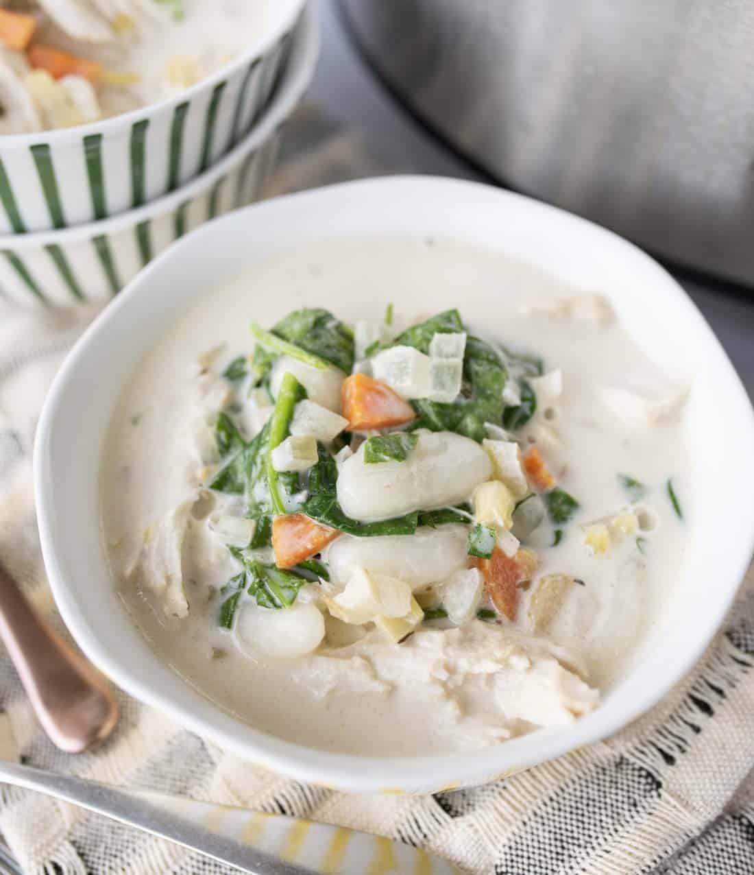 A bowl of creamy soup filled with gnocchi, shredded chicken, spinach, carrots, and onions, with another bowl of soup in the background on a striped cloth.