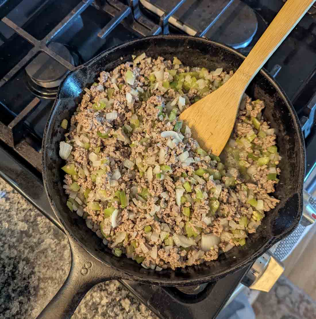 A cast iron skillet on a stove contains cooked ground beef mixed with chopped onions and green peppers. A wooden spoon is resting in the skillet, stirring the mixture.