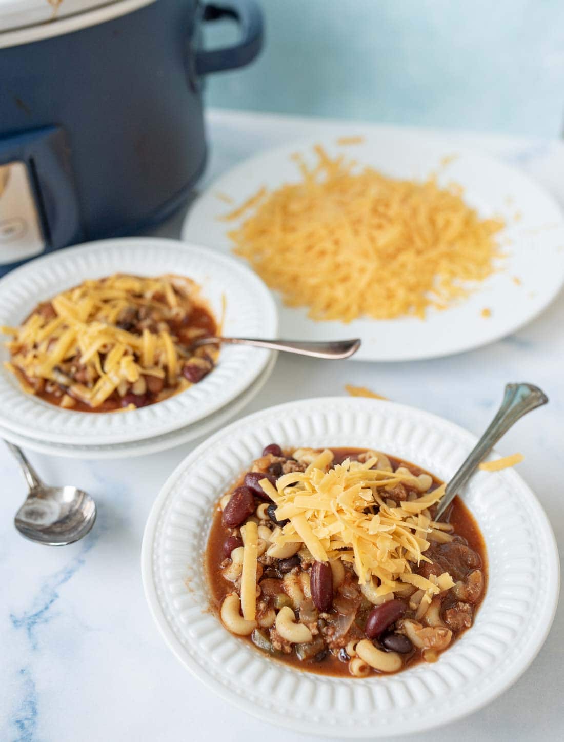 Two white bowls of slow cooker chili mac with chili, macaroni, and kidney beans, topped with shredded cheddar cheese, on a white surface. A plate of extra grated cheese and a slow cooker are in the background.