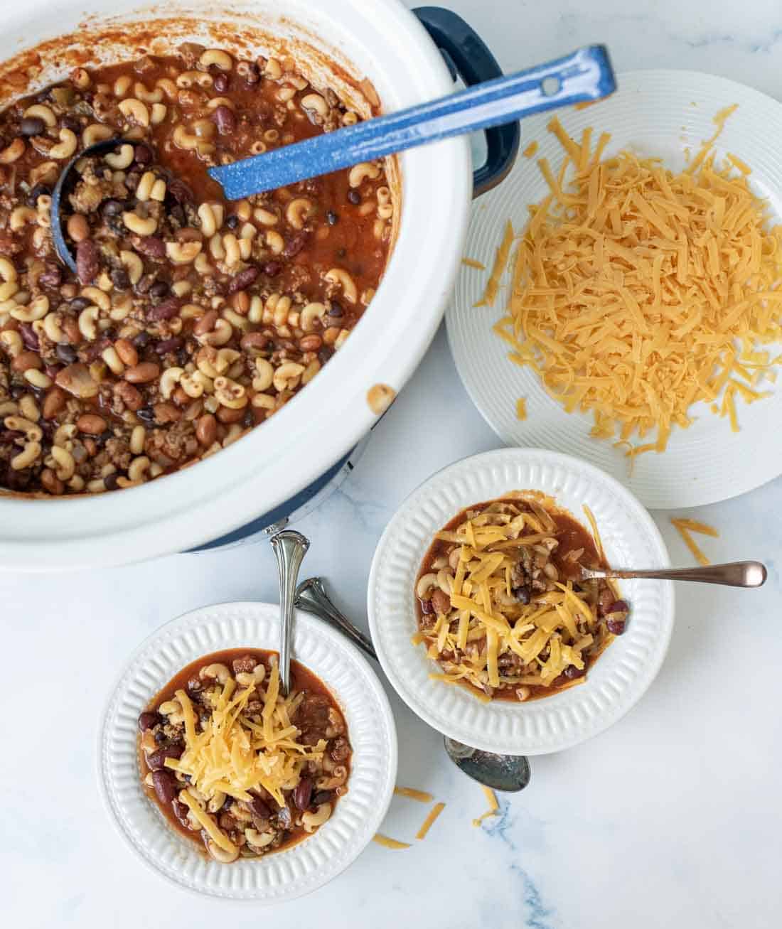 A crockpot filled with chili containing macaroni, beans, and ground meat sits beside a plate of shredded cheddar cheese and two bowls of chili topped with cheese on a white surface. Spoons are placed in the bowls.