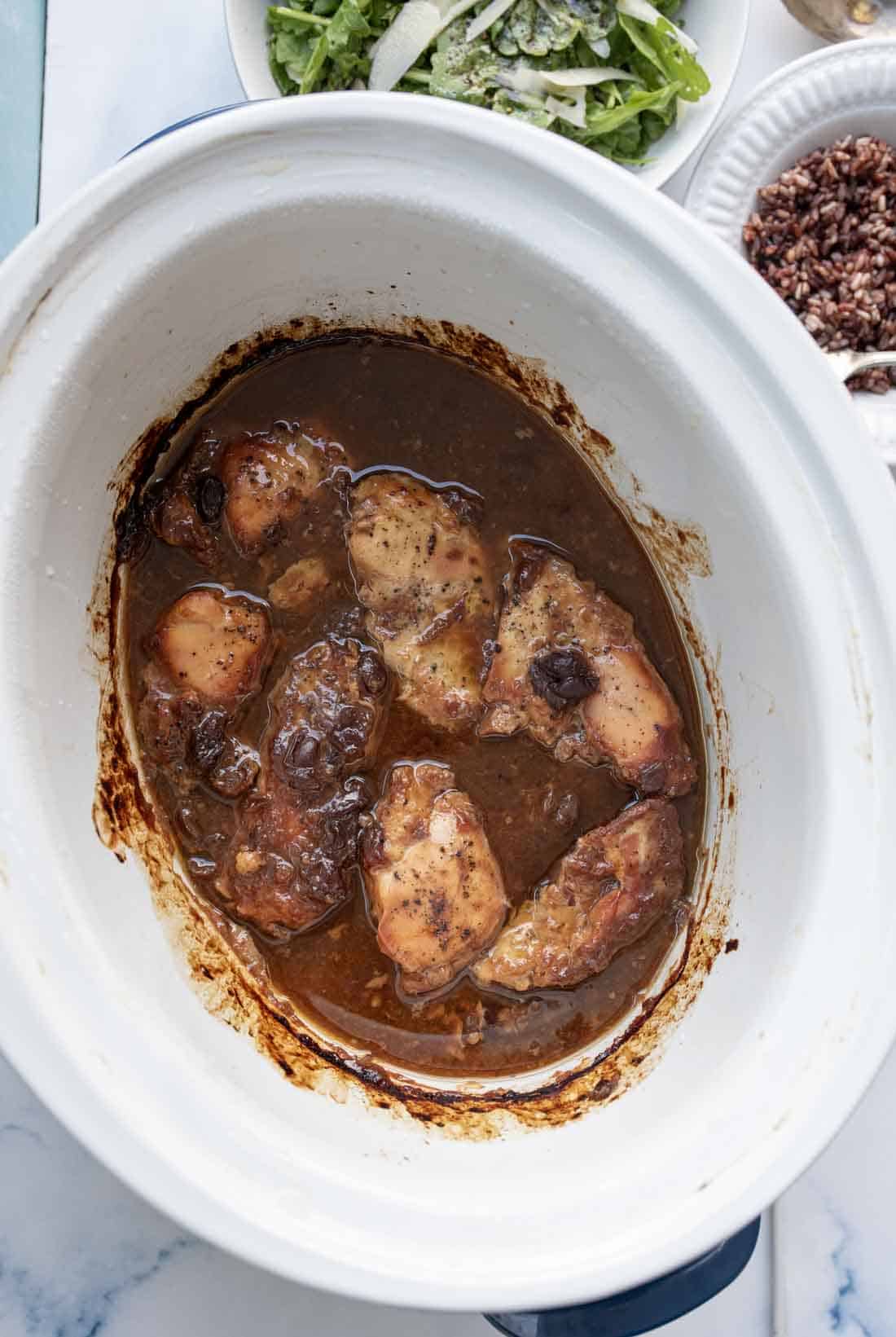 Chicken thighs cooked in brown sauce inside a white slow cooker, with a plate of salad and a bowl of rice partially visible in the background.
