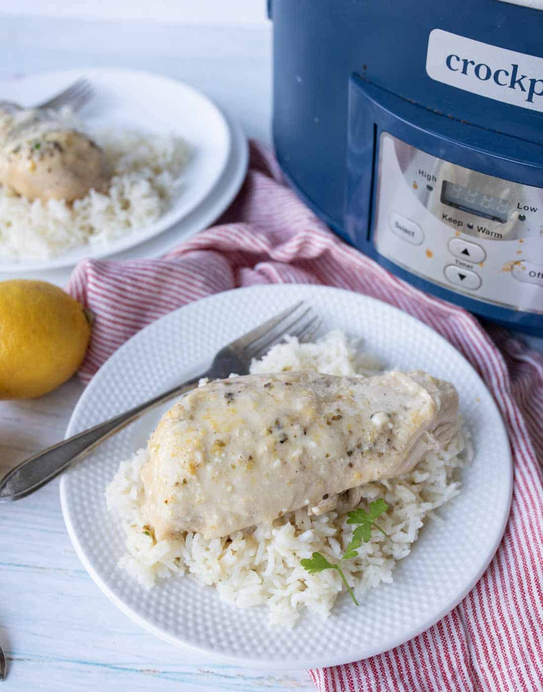 A plate of white rice topped with a piece of cooked chicken, garnished with parsley, sits beside a fork and knife. A slow cooker, lemon, and red-striped cloth are in the background.