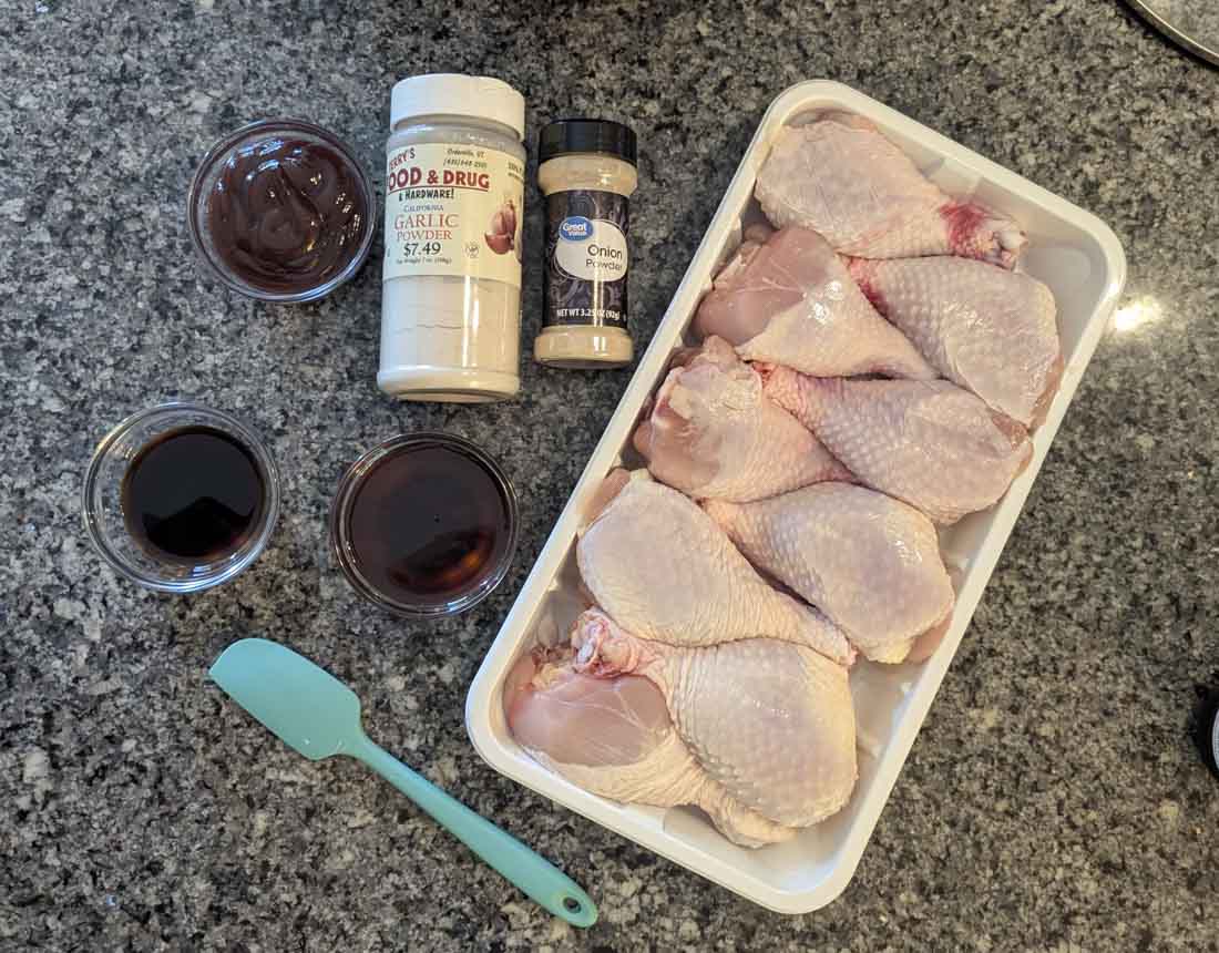 Raw chicken drumsticks in a plastic tray, with small bowls of barbecue sauce and soy sauce, a teal spatula, and containers of garlic powder and onion powder on a granite countertop.