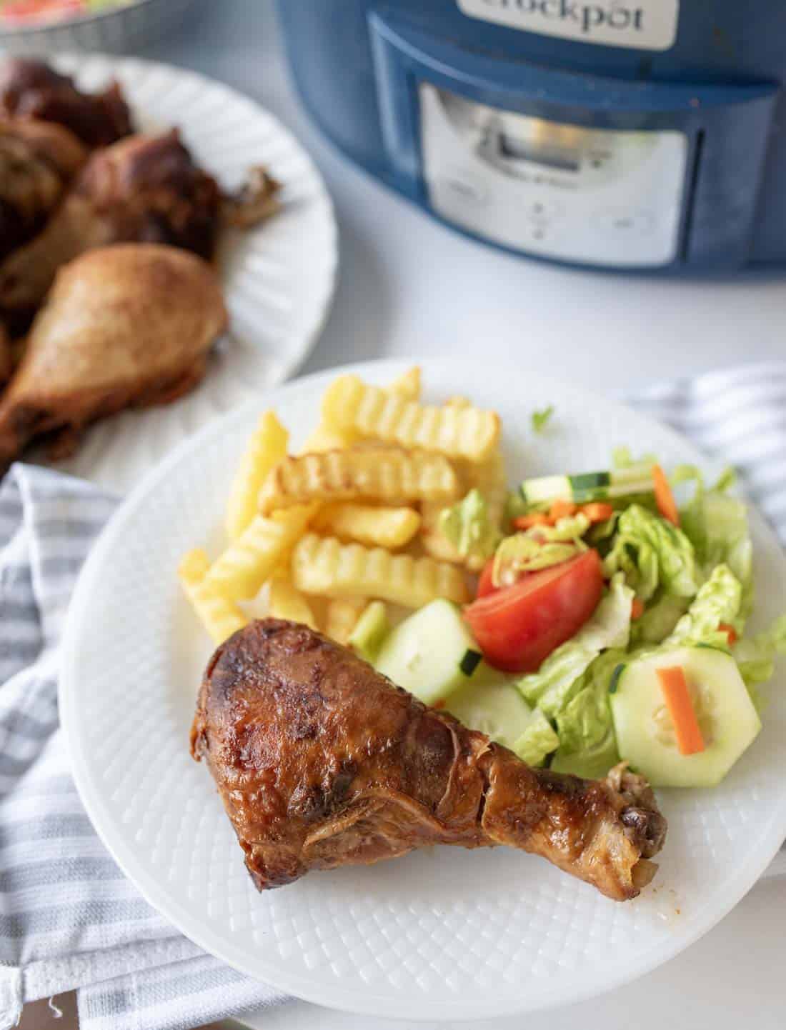 A white plate with a cooked chicken drumstick, crinkle-cut fries, and a fresh salad with lettuce, tomato, cucumber, and carrot. In the background, a slow cooker and another plate with more chicken are visible.