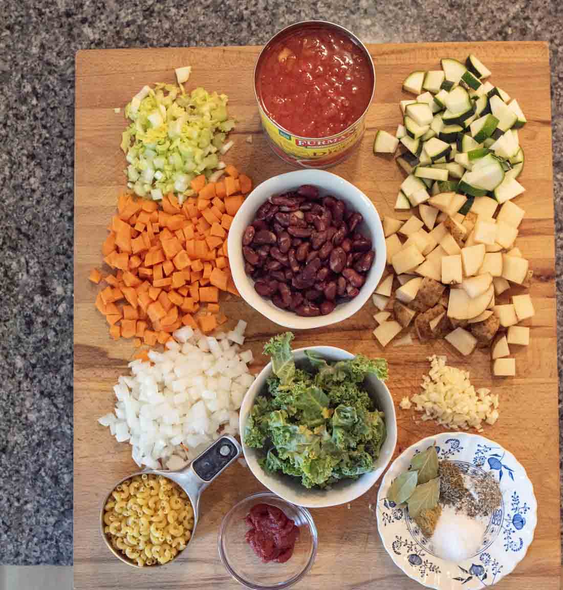 A wooden cutting board displays chopped celery, carrots, zucchini, potatoes, onions, garlic, kale, kidney beans, a can of diced tomatoes, elbow macaroni, tomato paste, and small bowls of spices and herbs.