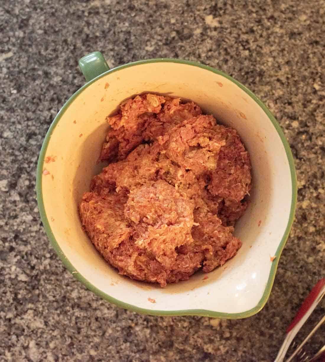 A green mixing bowl filled with seasoned raw ground meat mixture sits on a speckled kitchen countertop.