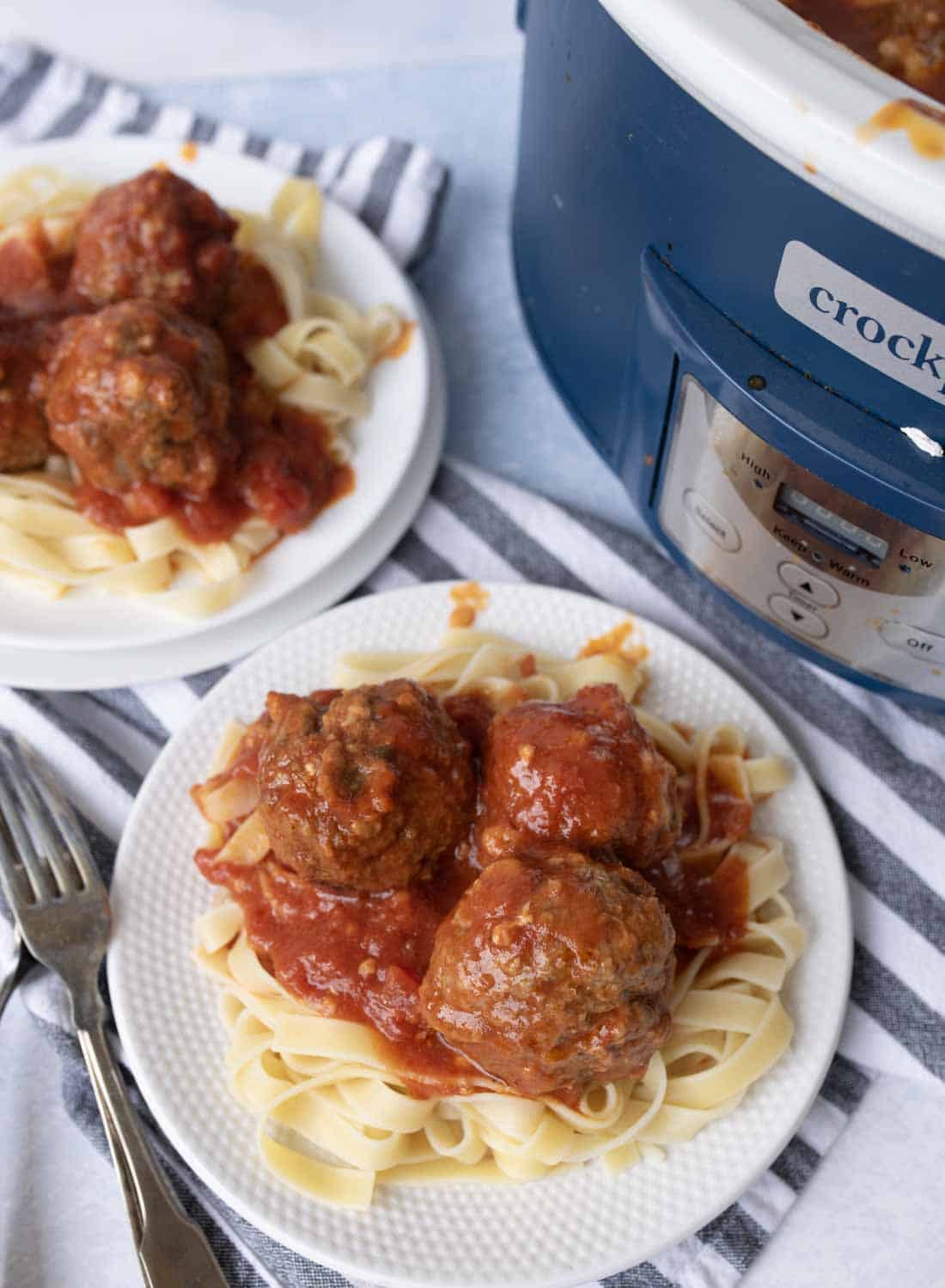 Plates of tagliatelle pasta topped with meatballs in tomato sauce are set next to a blue slow cooker on a striped cloth, with forks beside the dishes.
