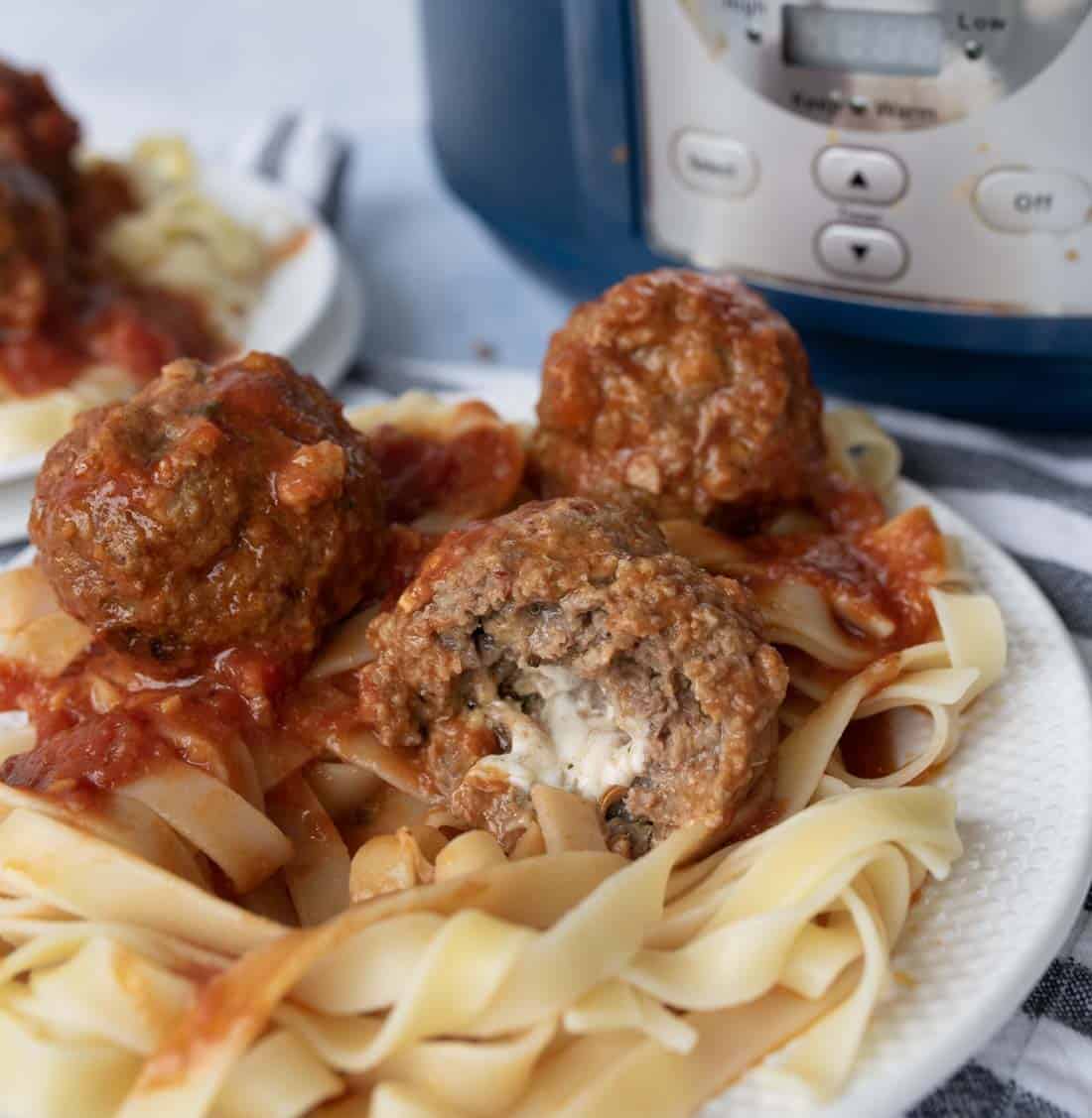 A plate of fettuccine noodles topped with tomato sauce and meatballs, one of which is cut open to show a cheese-filled center. An electric pressure cooker is visible in the background.
