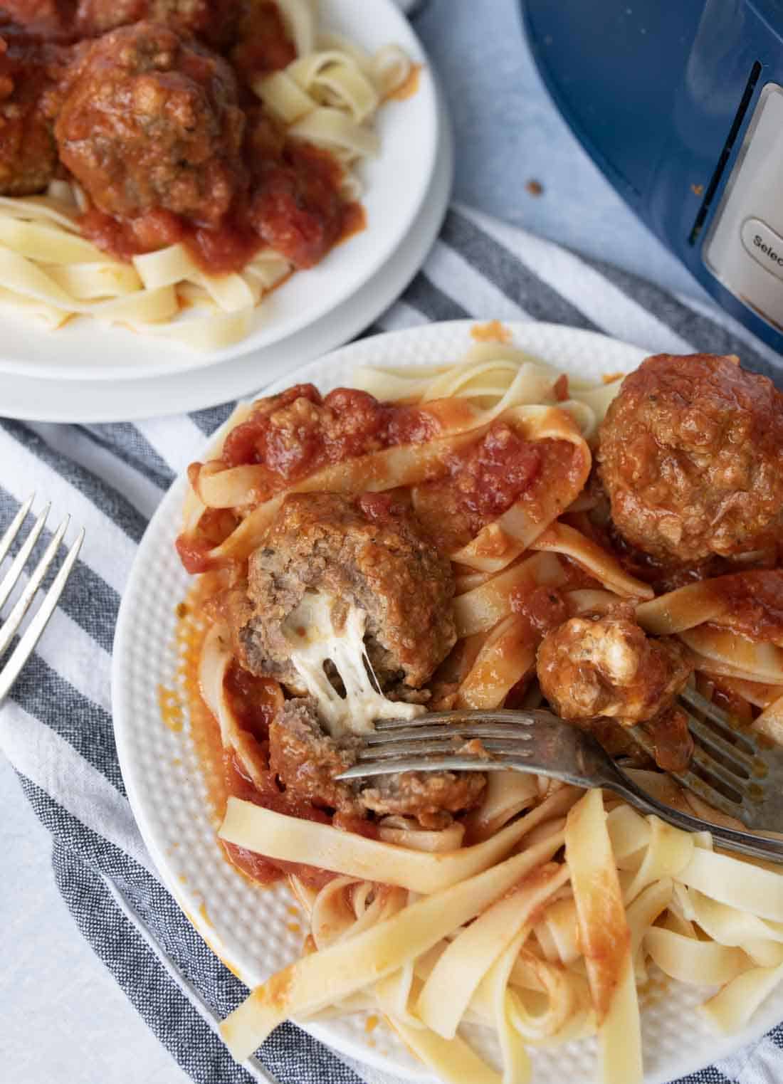 A plate of fettuccine pasta topped with tomato sauce and mozzarella stuffed meatballs, with a fork cutting into a meatball; another plate with the same meal is in the background.