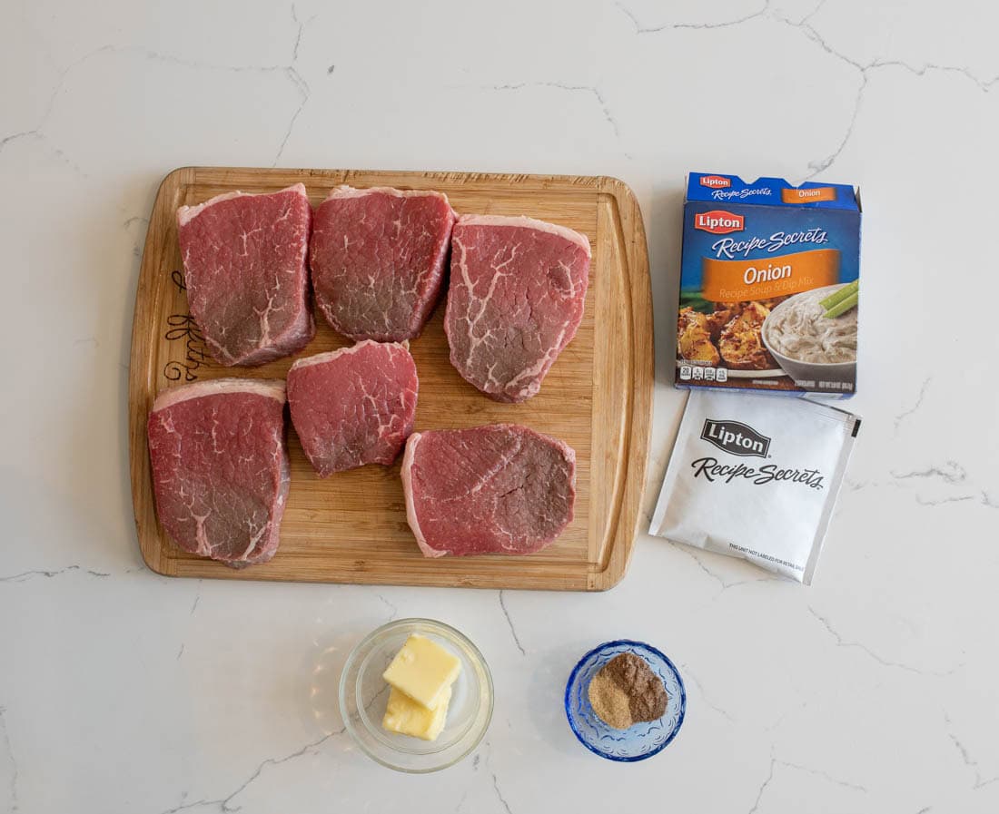 Six raw beef steaks on a wooden cutting board next to a box and packet of Lipton Onion Soup mix, a bowl of butter, and a small bowl of dry seasoning on a white countertop.