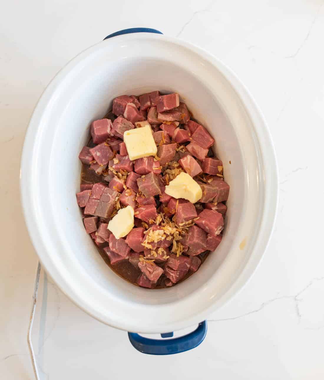 Overhead view of a slow cooker filled with raw cubed beef, topped with chopped onions and several pieces of butter, on a white countertop.