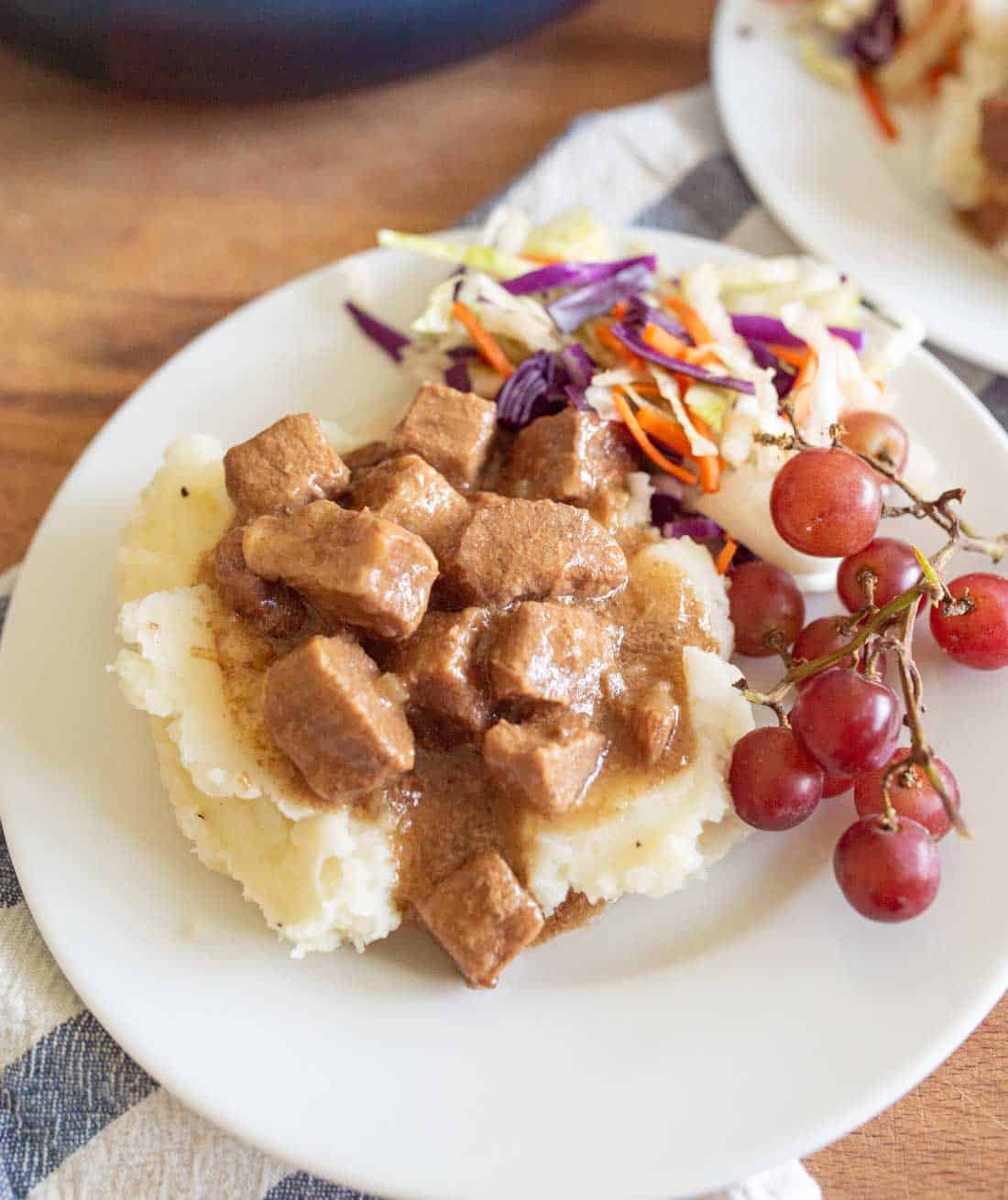 A plate of mashed potatoes topped with slow cooker steak bites, served with coleslaw and a small bunch of red grapes on the side.