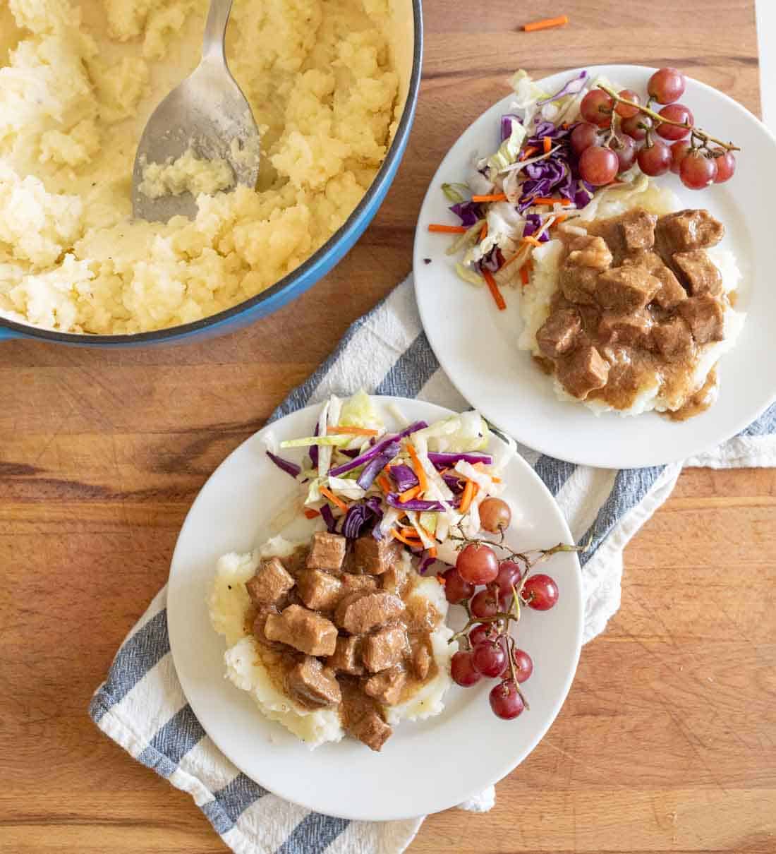 Two plates with mashed potatoes topped with brown gravy and meat cubes, served with a grape bunch and colorful coleslaw. A pot of mashed potatoes and a serving spoon sit nearby on a wooden surface with a striped towel.