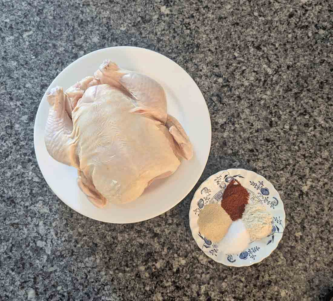 A whole raw chicken on a white plate sits next to a small plate with piles of various powdered spices, all on a gray speckled countertop.