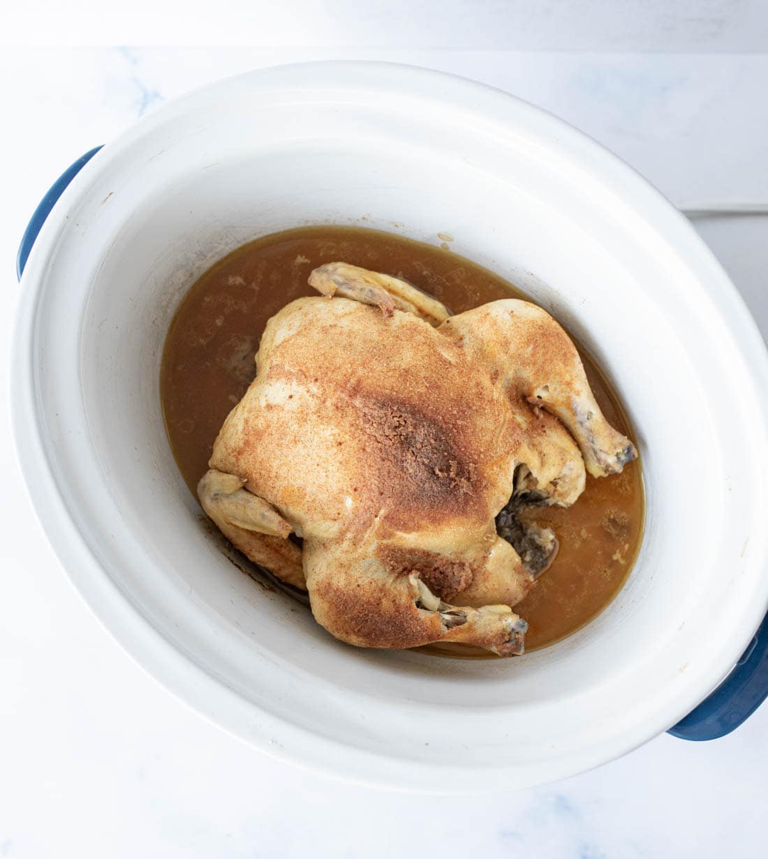 A whole cooked chicken with seasoned skin sits in a white slow cooker, surrounded by brown cooking juices.