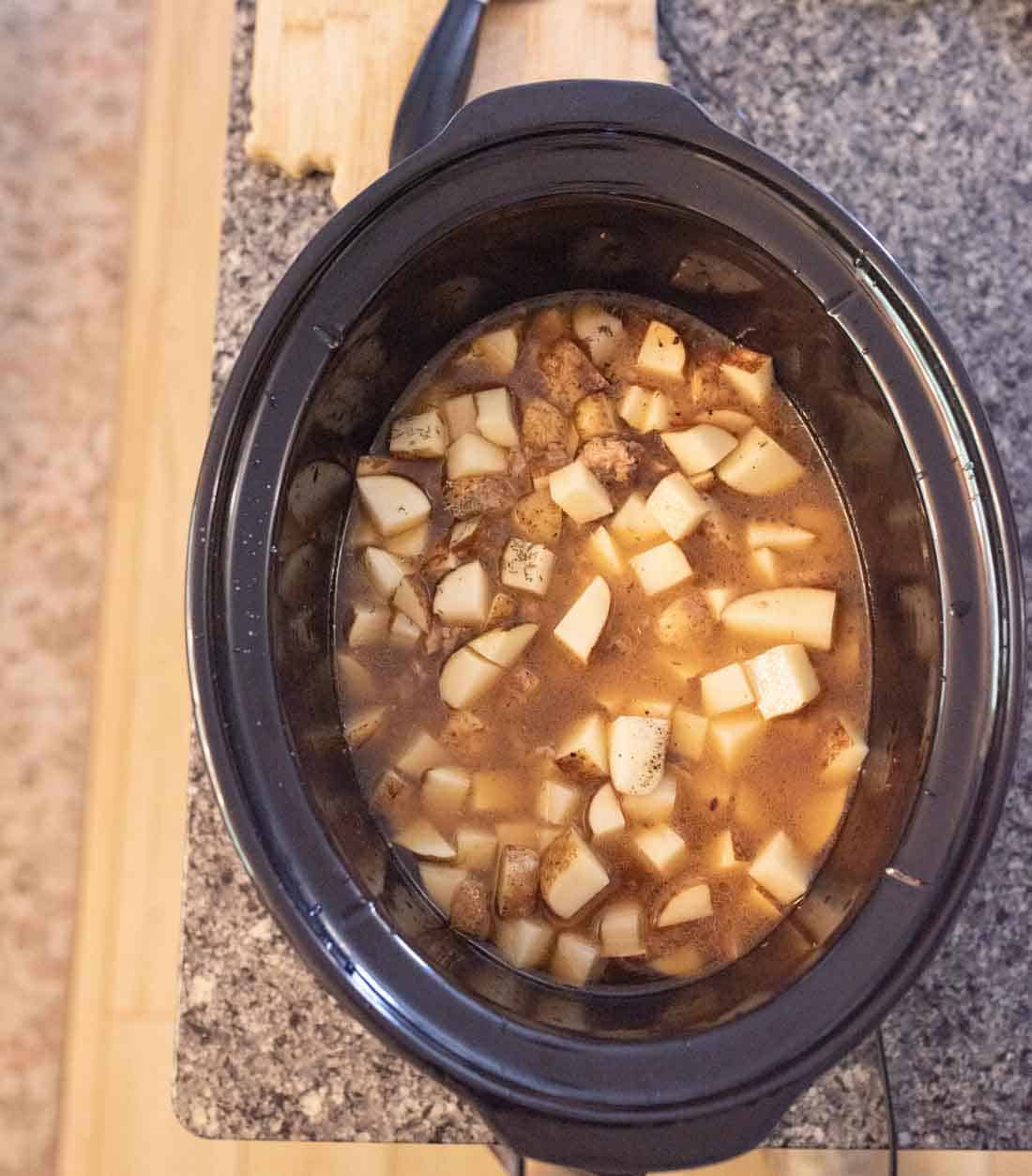 A slow cooker filled with a mixture of diced potatoes, beef chunks, and broth sits on a kitchen countertop next to a wooden cutting board.