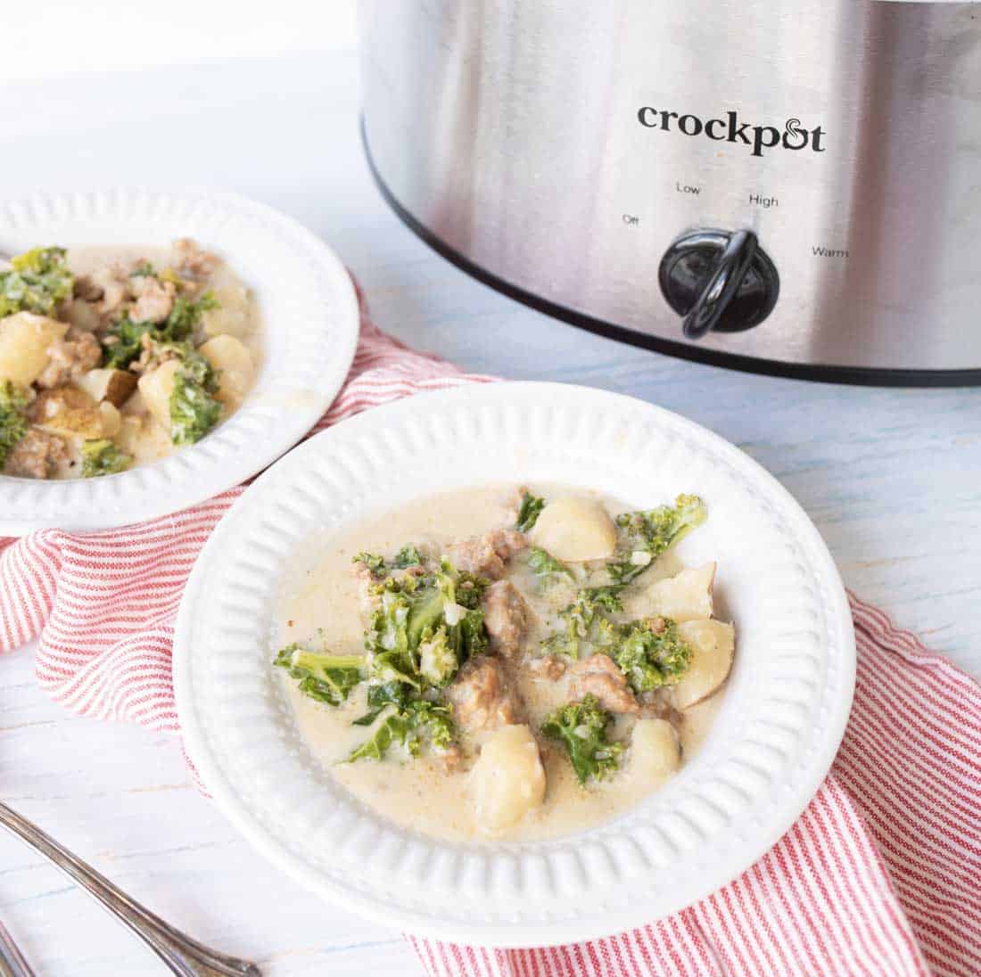 Two white bowls filled with creamy sausage potato soup containing sausage, potatoes, and kale sit on red striped napkins in front of a silver Crockpot slow cooker.