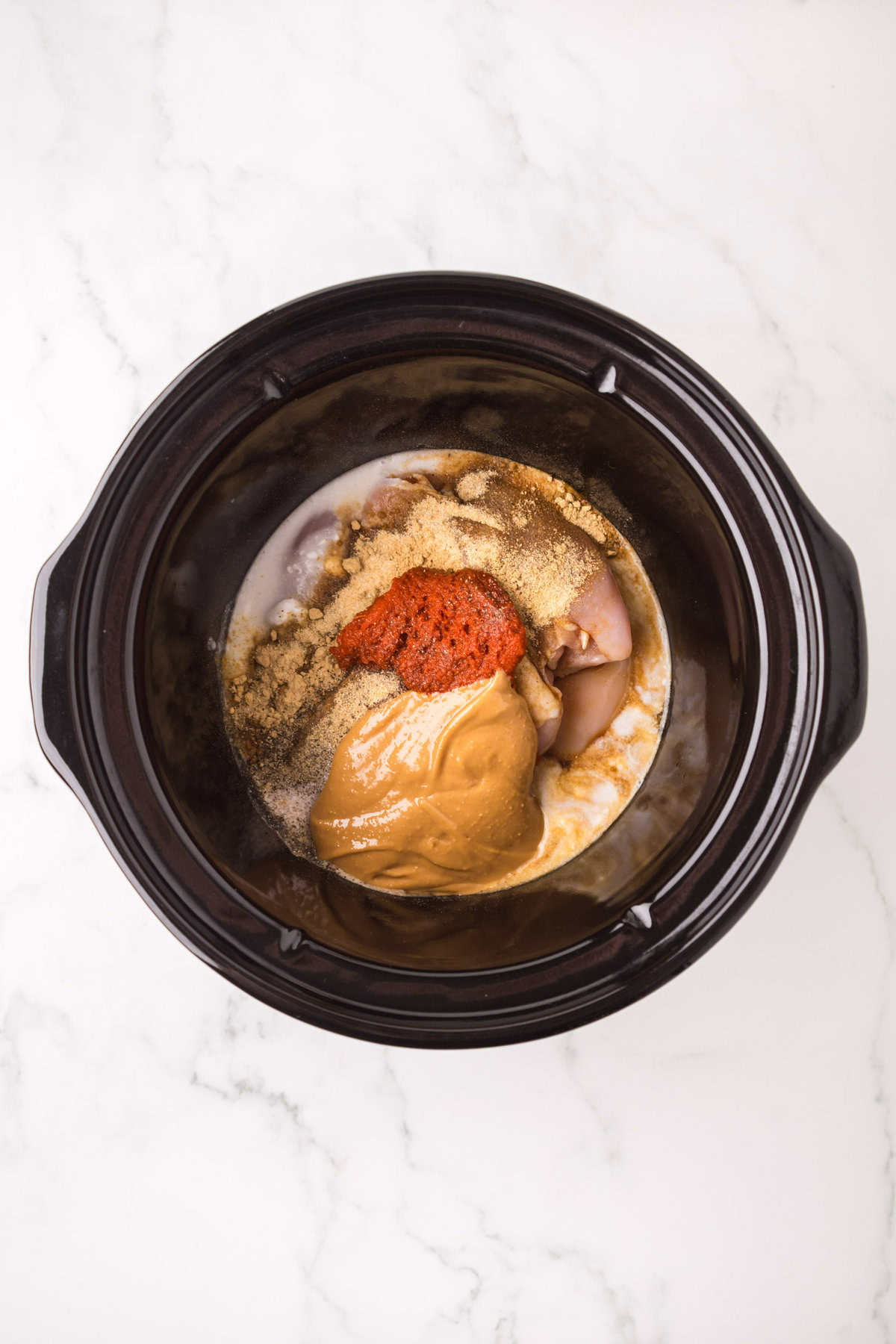 A slow cooker filled with raw chicken, peanut butter, spices, and seasonings for an easy Thai chicken satay recipe, ready to be mixed and cooked, placed on a white marble countertop.