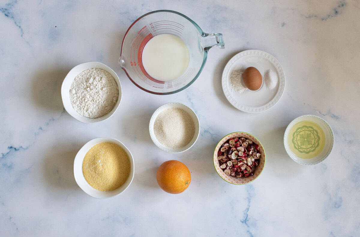 Bowls of flour, cornmeal, sugar, dried cranberries, and oil, along with a measuring cup of milk, a whole orange, and a small plate holding an egg and two piles of powder, all arranged on a marble surface.