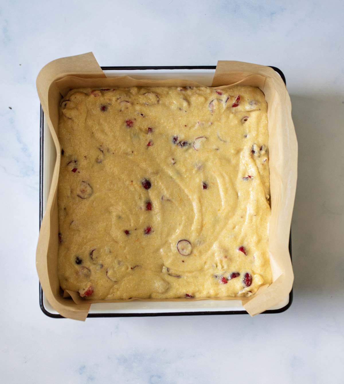 A square baking pan lined with parchment paper holds unbaked batter with visible pieces of fruit or nuts, set on a light marble surface.