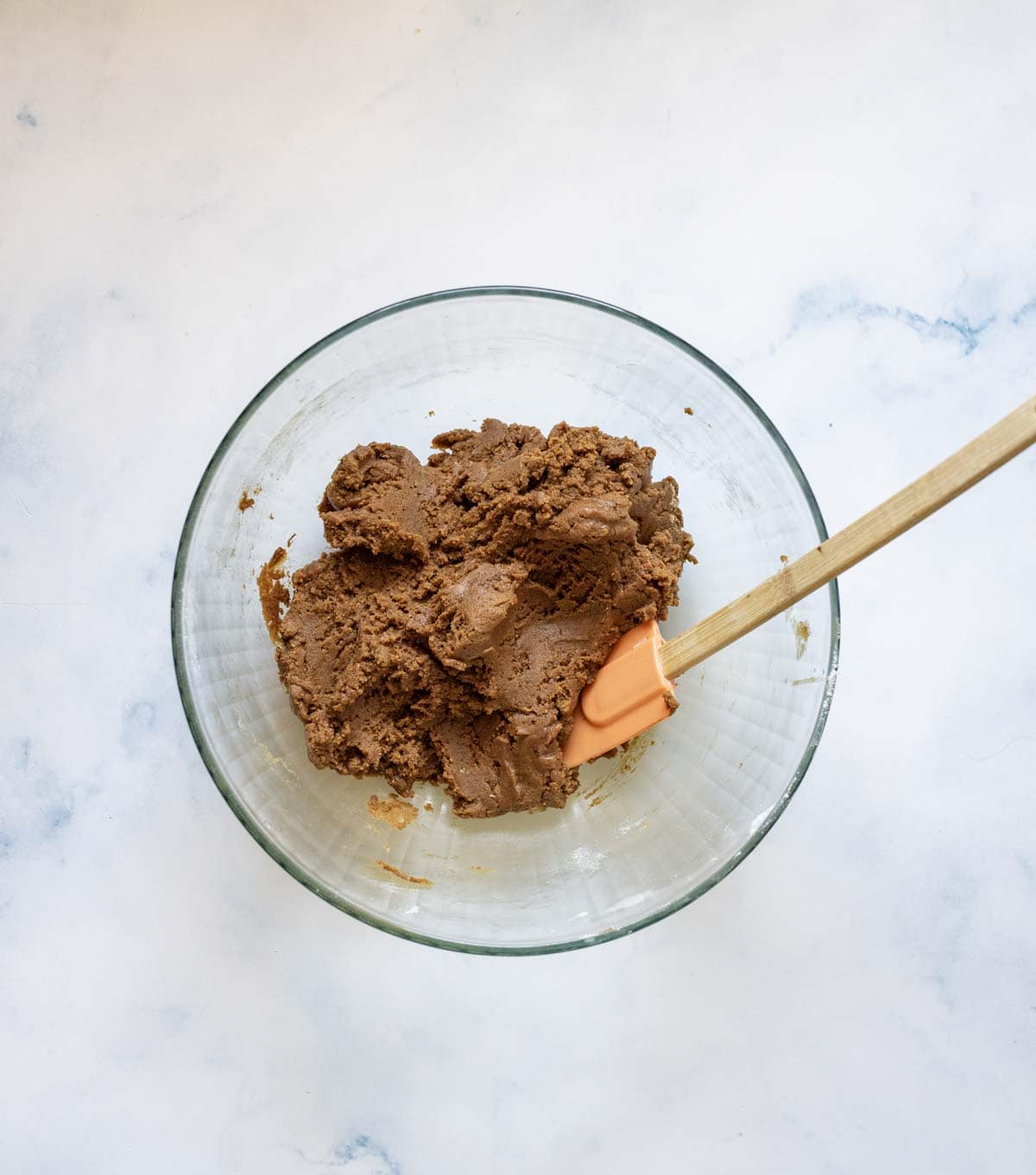 A glass bowl filled with chocolate cookie dough sits on a white surface. An orange spatula with a wooden handle rests in the dough.