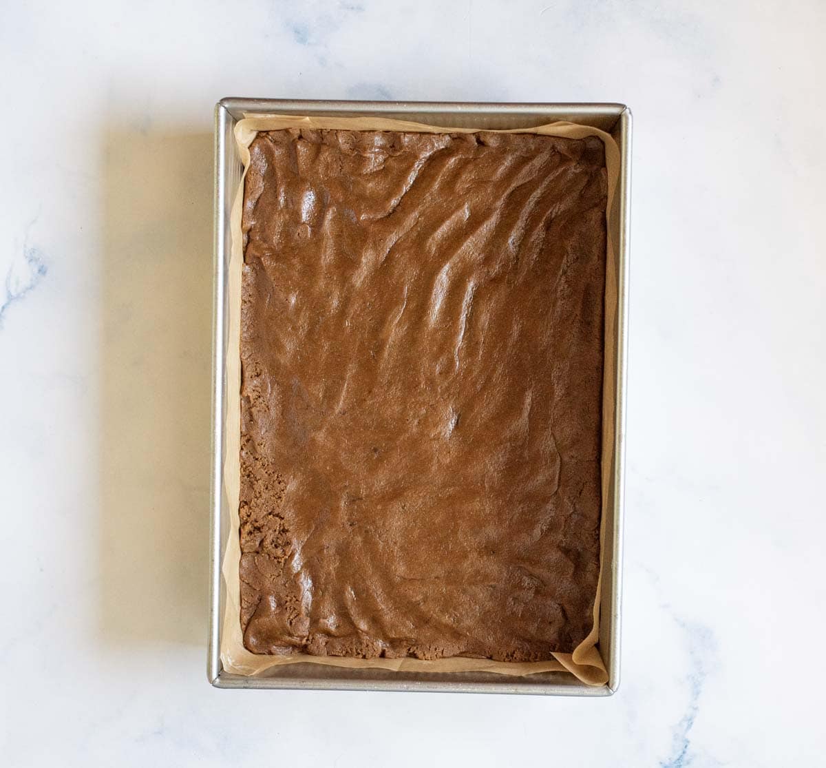 A rectangular pan lined with parchment paper, filled with a large uncut sheet of baked brownies on a white marble surface.