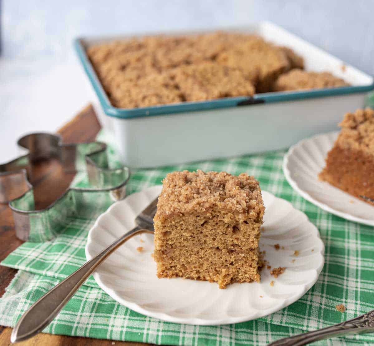 A slice of crumb-topped coffee cake sits on a white plate with a fork, placed on a green plaid cloth. In the background is a baking dish with more cake and a metal cookie cutter.