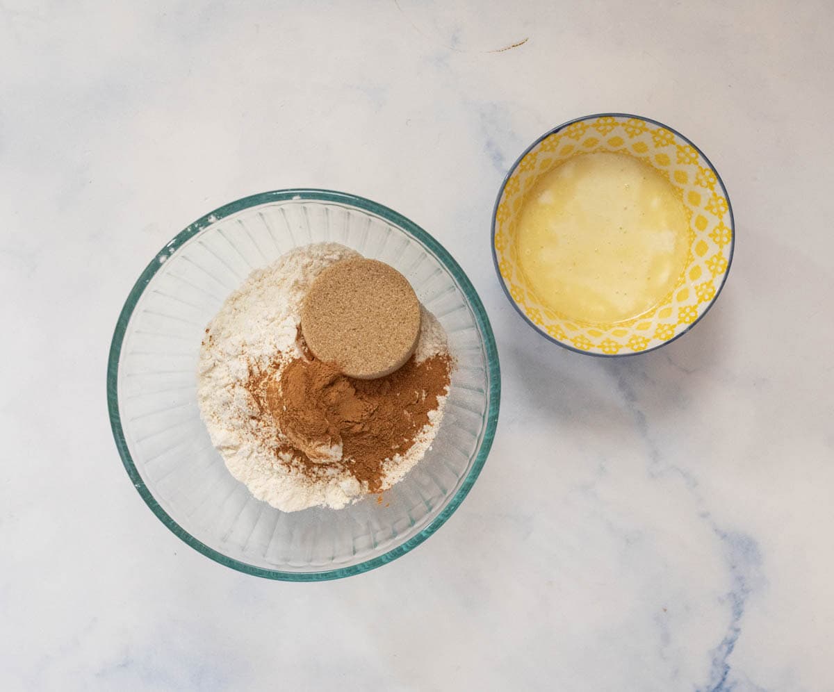 A glass bowl with flour, brown sugar, and cinnamon beside a yellow-patterned bowl containing melted butter, both on a light marble surface.