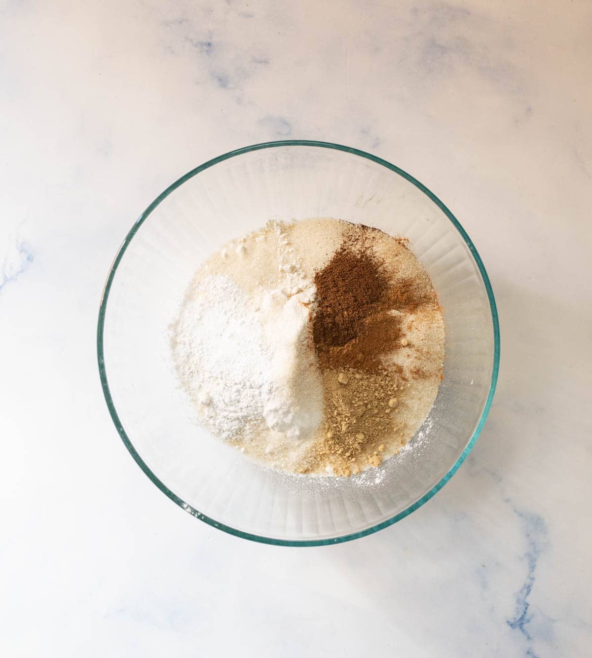 A glass bowl containing separate piles of dry baking ingredients, including flour, sugar, and ground spices, on a light marble surface.