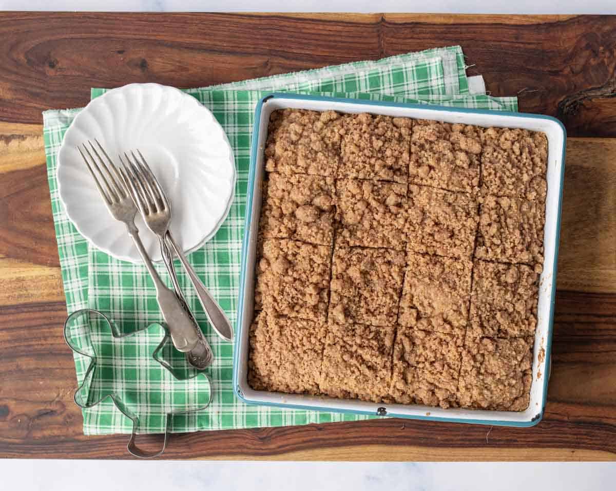 A pan of crumb-topped gingerbread coffee cake, cut into squares, sits on a green plaid napkin with two white plates and three forks on the side, atop a wooden surface. Two metal cookie cutters are also visible.