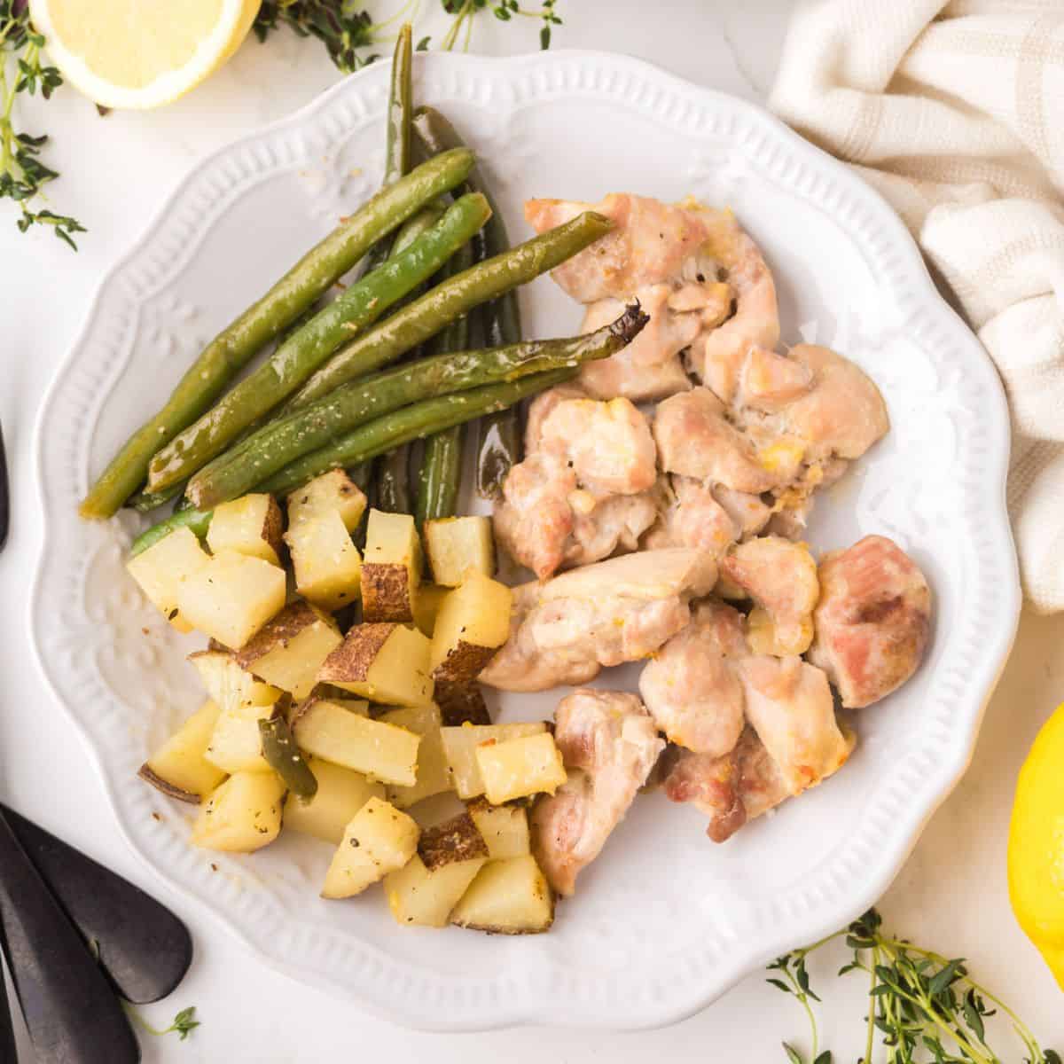 A white plate with roasted potatoes, green beans, and pieces of baked chicken, garnished with fresh herbs. A lemon wedge and a white cloth are nearby on the table.