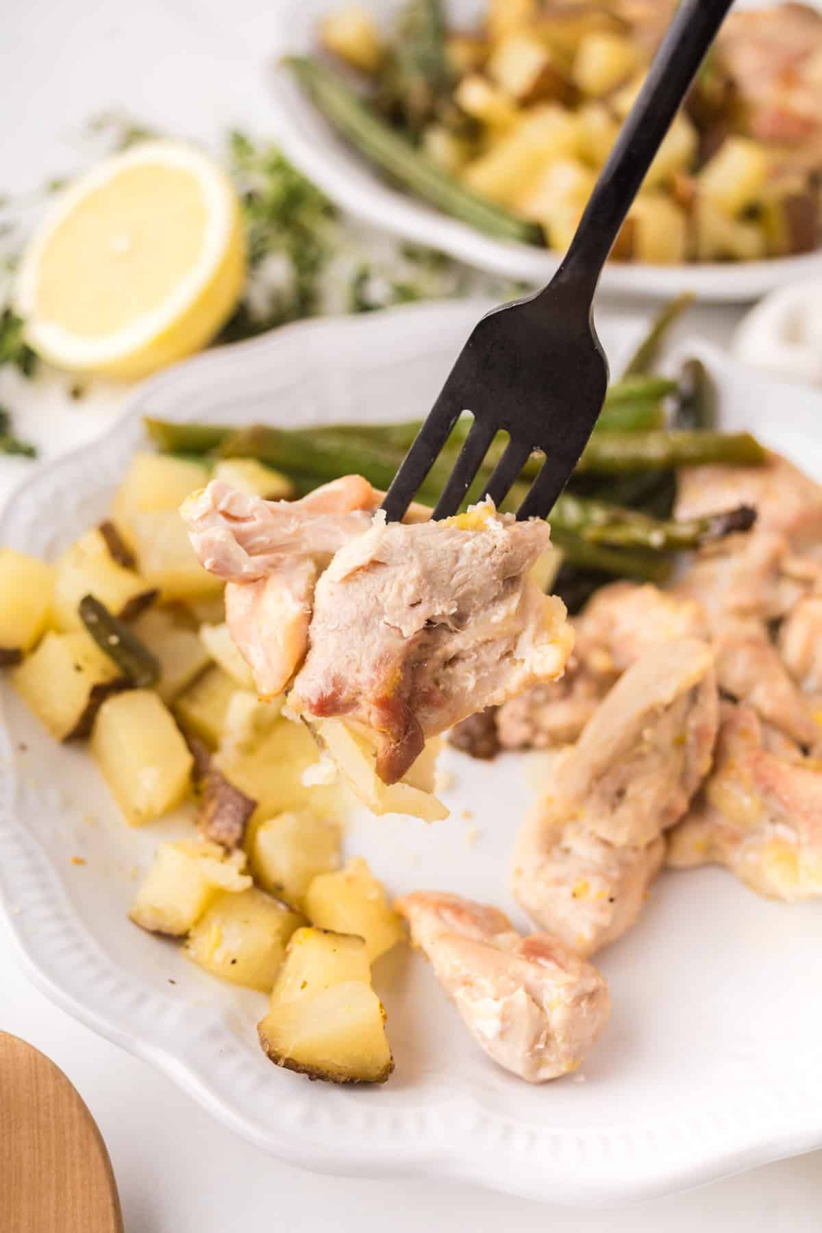 A fork holds a bite of cooked chicken over a white plate with roasted potatoes and green beans. A lemon slice and fresh herbs are in the background.