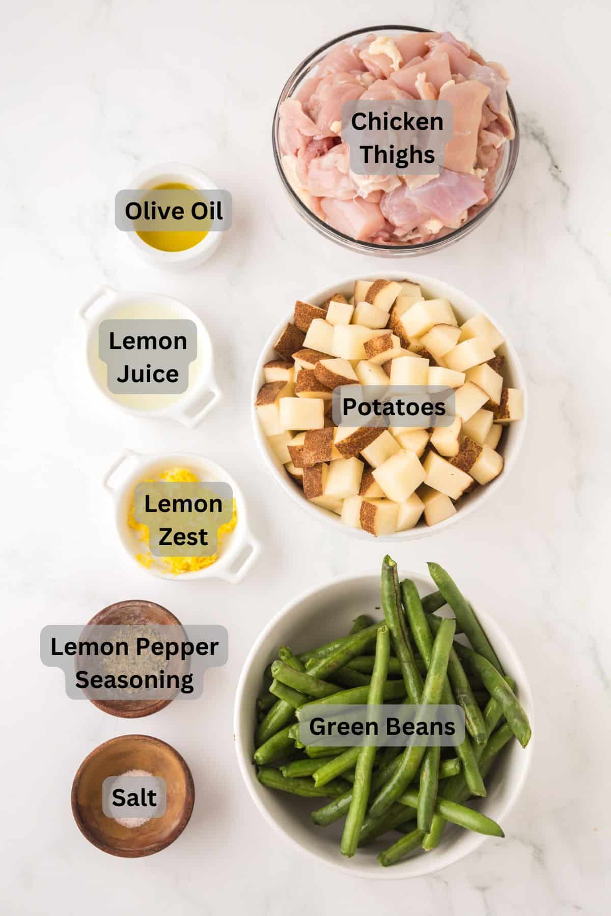 Top-down view of labeled ingredients on a white surface: chicken thighs, diced potatoes, green beans, olive oil, lemon juice, lemon zest, lemon pepper seasoning, and salt in separate bowls.
