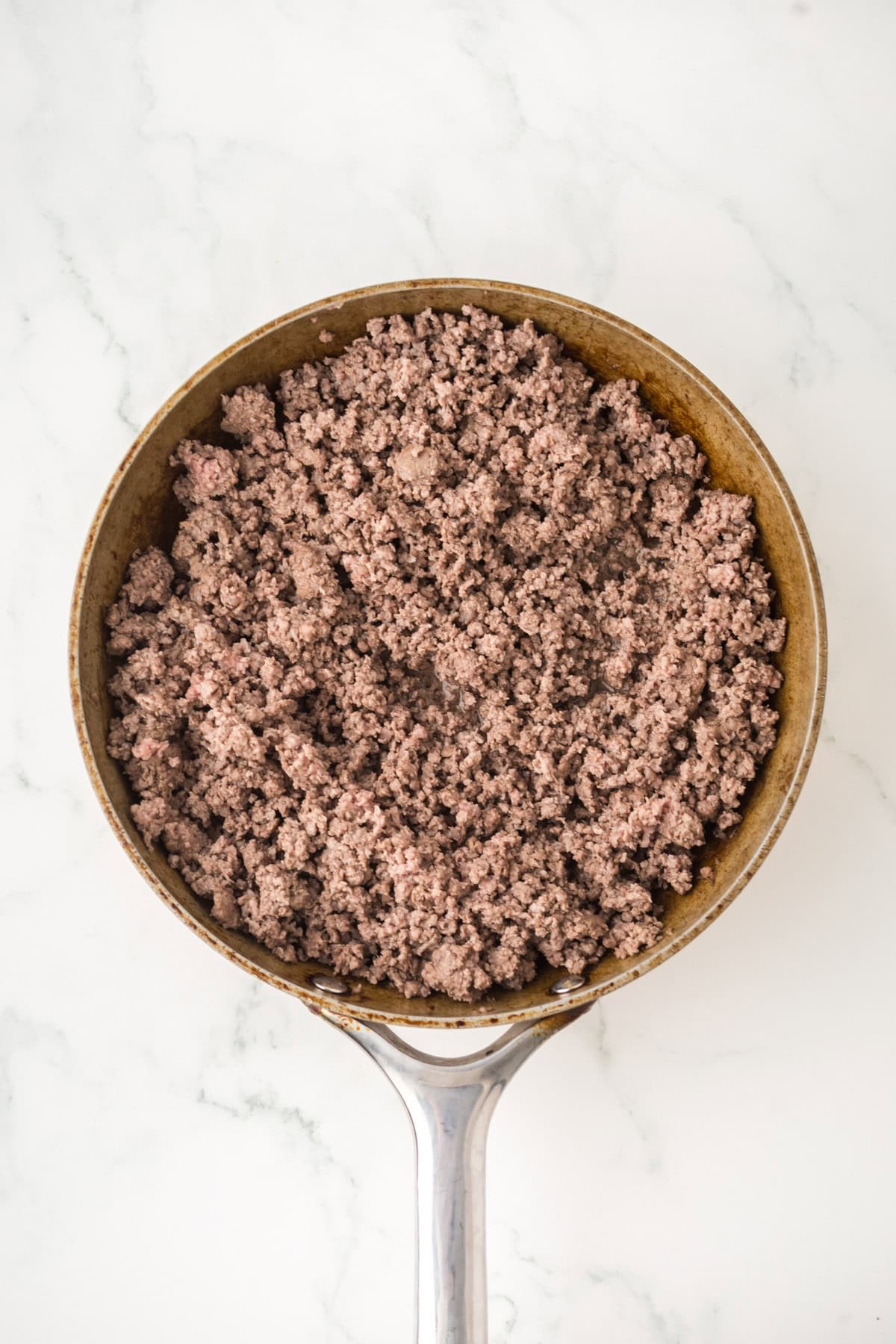 A frying pan filled with cooked ground beef, perfect for making crock pot sloppy joes, sits on a white marble surface.