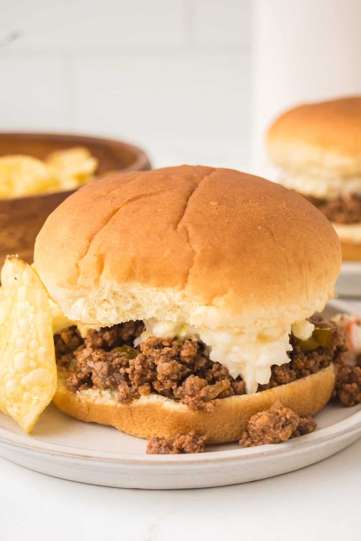 Crock pot sloppy joes sandwich with ground beef filling and a dollop of coleslaw is served on a plate with potato chips. Another crock pot sloppy joes sandwich and more chips are visible in the background.