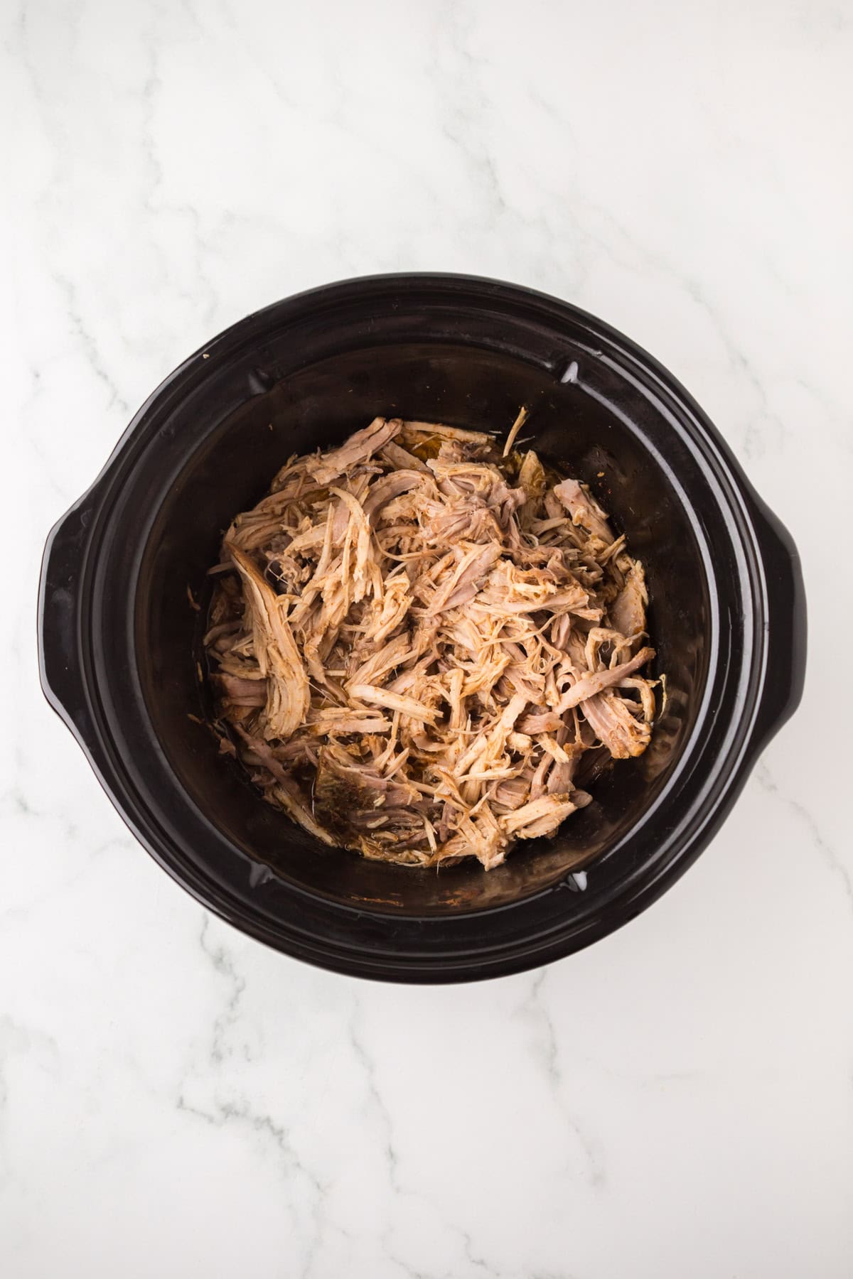 Shredded pork in a black slow cooker sits on a white marble surface, ready to be the star ingredient for delicious pulled pork nachos.