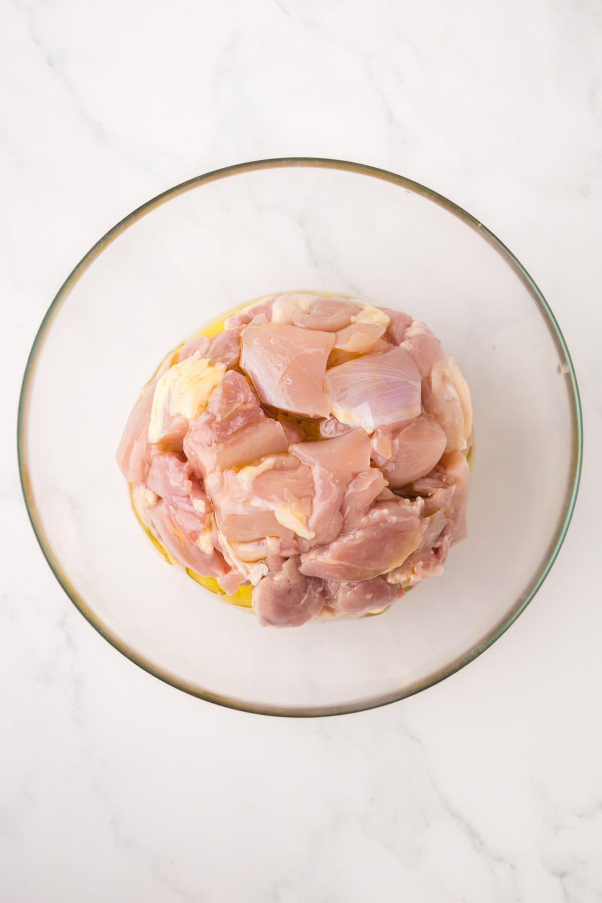 Raw, cubed pieces of chicken placed in a clear glass bowl on a white marble surface.