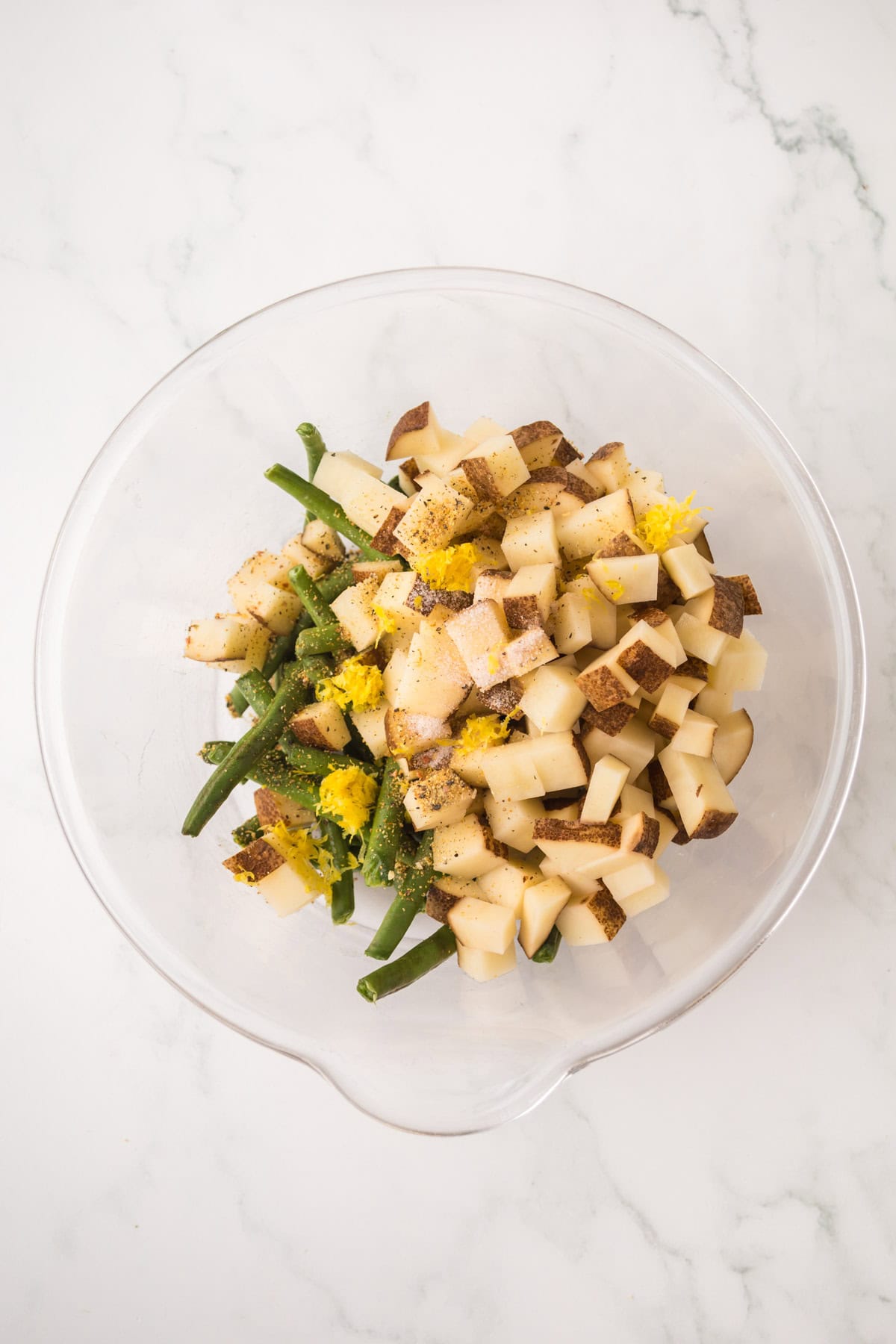 A glass mixing bowl containing chopped potatoes, green beans, lemon zest, and ground black pepper, all sitting on a white marble surface.