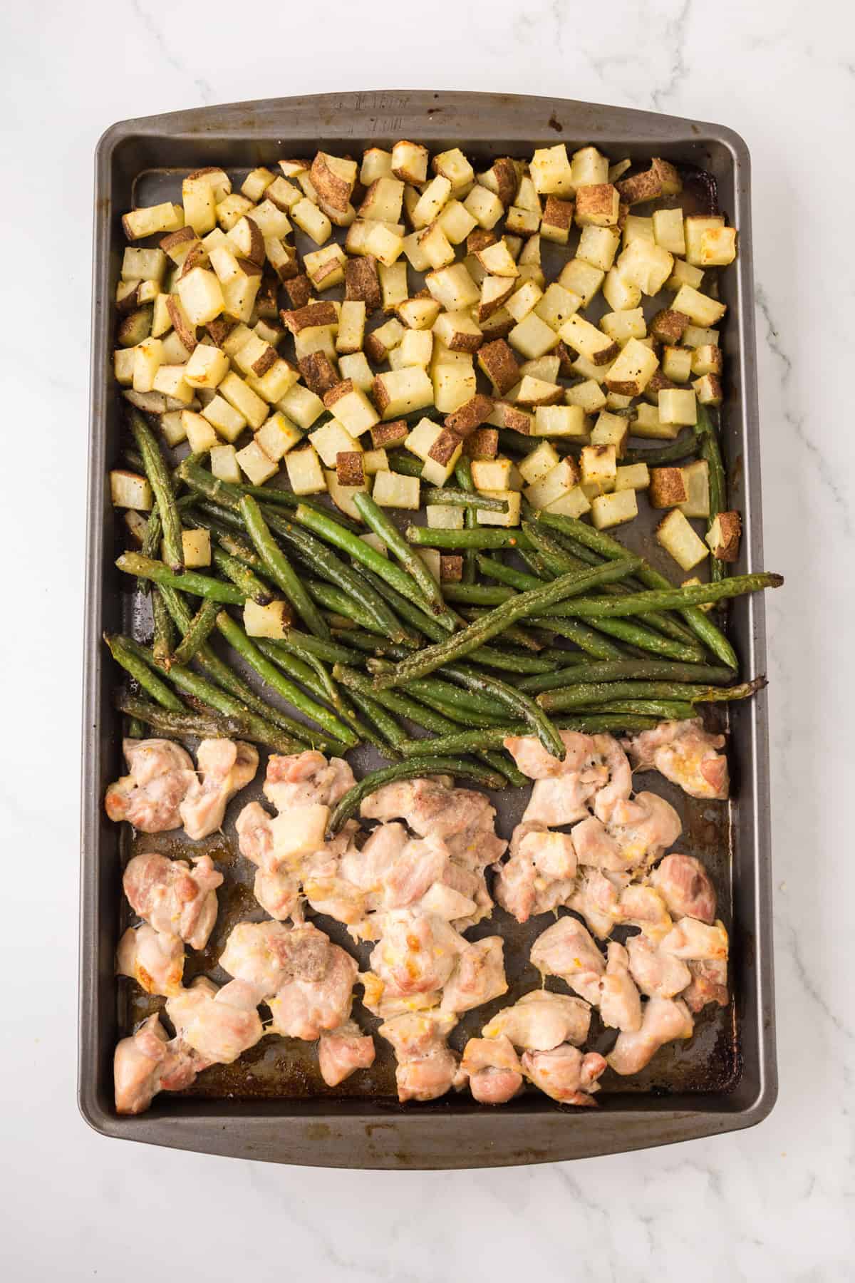 A sheet pan with roasted diced potatoes, green beans, and pieces of cooked chicken thigh arranged in sections on a marble countertop.