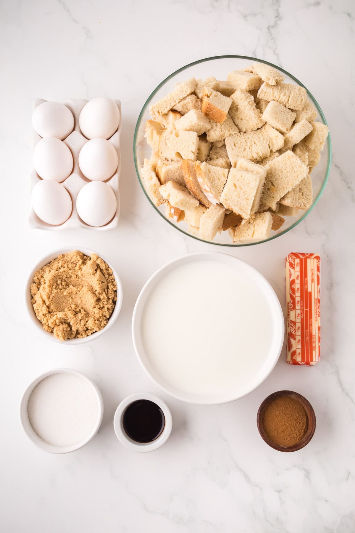 A top-down view of ingredients for bread pudding: a bowl of cubed bread, a carton of eggs, brown sugar, white sugar, vanilla extract, cinnamon, a stick of butter, and a bowl of milk on a marble surface.