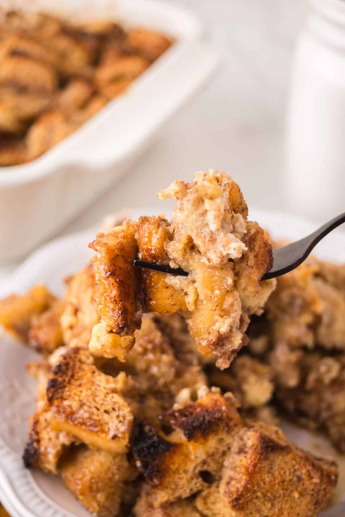 A close-up of a fork holding a bite of baked bread pudding with a golden-brown, cinnamon-coated crust. More bread pudding is served on a white plate and in a baking dish in the background.