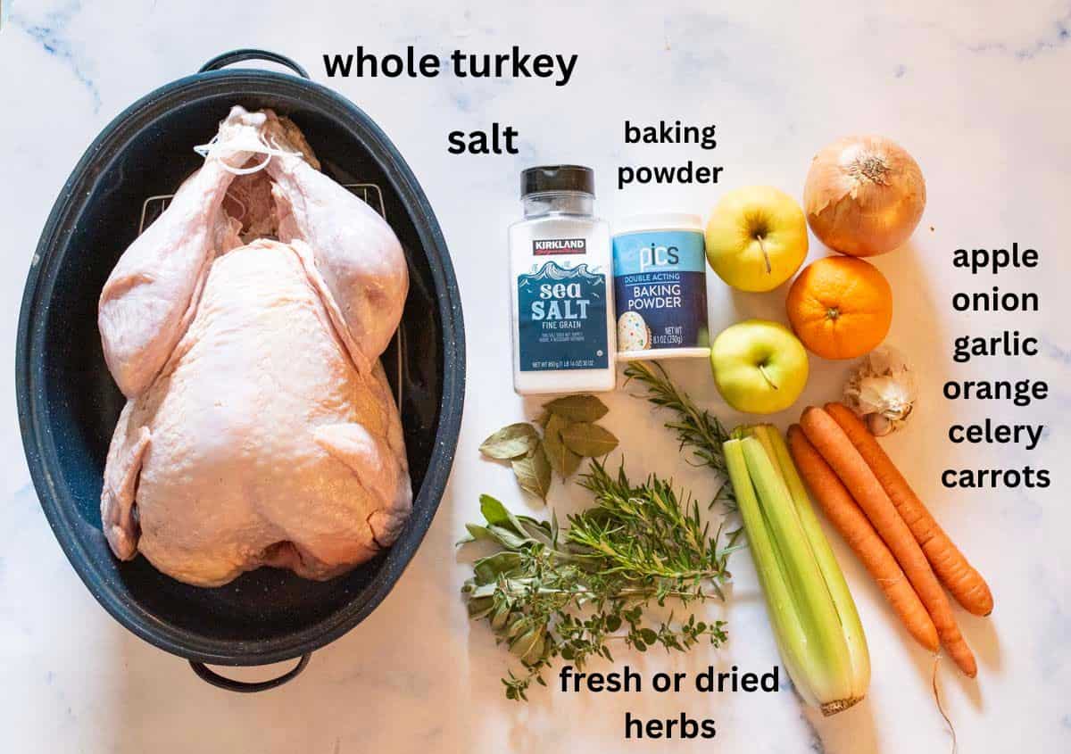 A whole raw turkey in a roasting pan sits next to salt, baking powder, fresh herbs, celery, carrots, an orange, garlic, an apple, and an onion, all arranged on a white surface with labels.