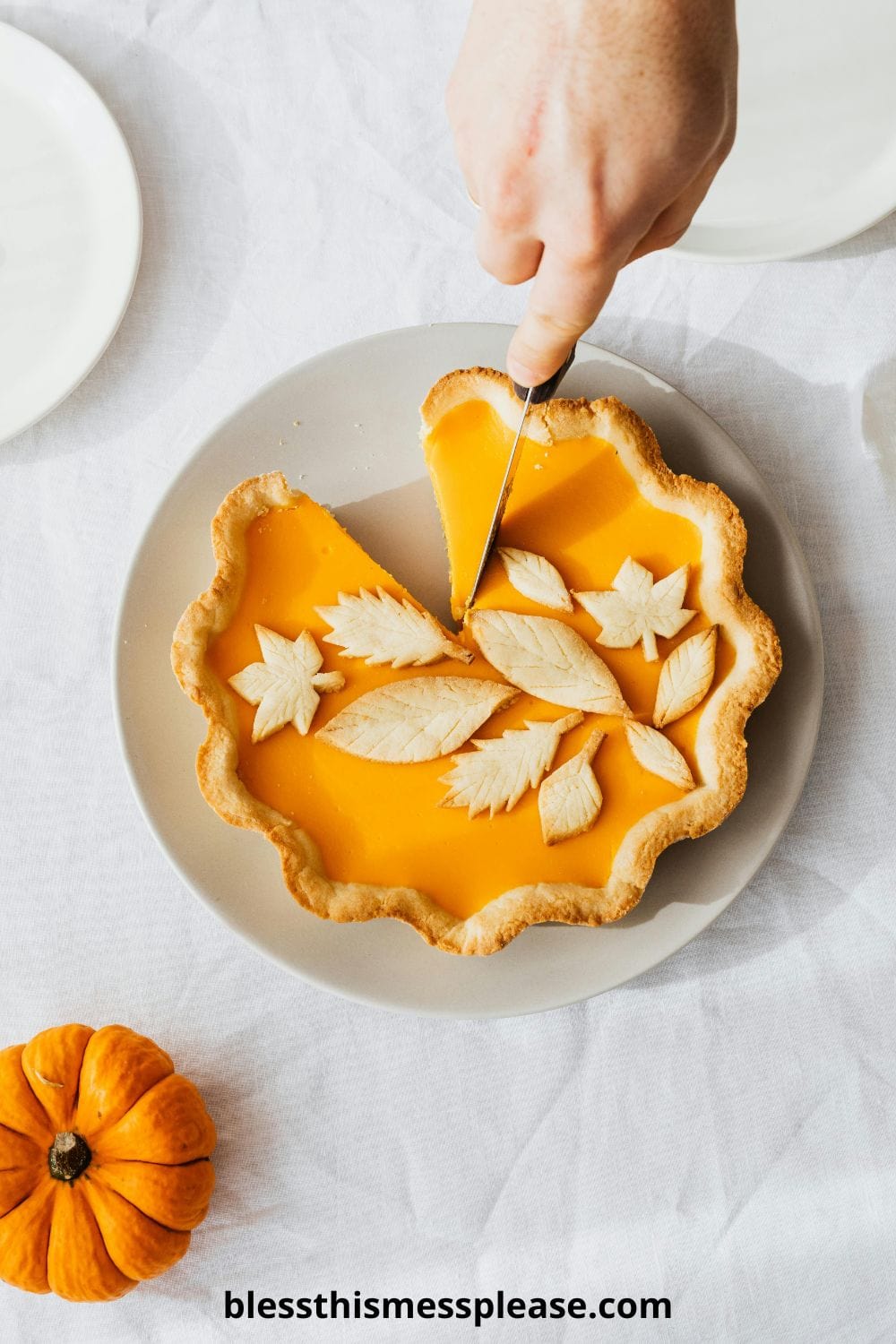 A hand slices a pumpkin pie with a decorative leaf-shaped crust on top, placed on a light gray plate. Showcasing how to make pie crust, the scene features a small pumpkin and an empty plate nearby on a white tablecloth. Website text appears below.