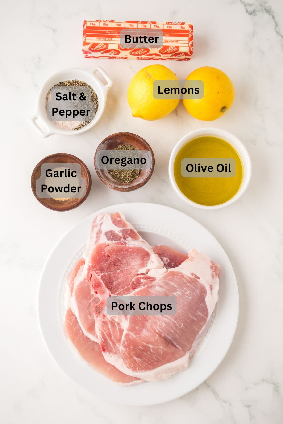 Overhead view of ingredients on a white surface for lemon pepper pork chops: pork chops on a plate, two lemons, butter, salt & pepper bowls, garlic powder, oregano, and olive oil. Each ingredient is clearly labeled.