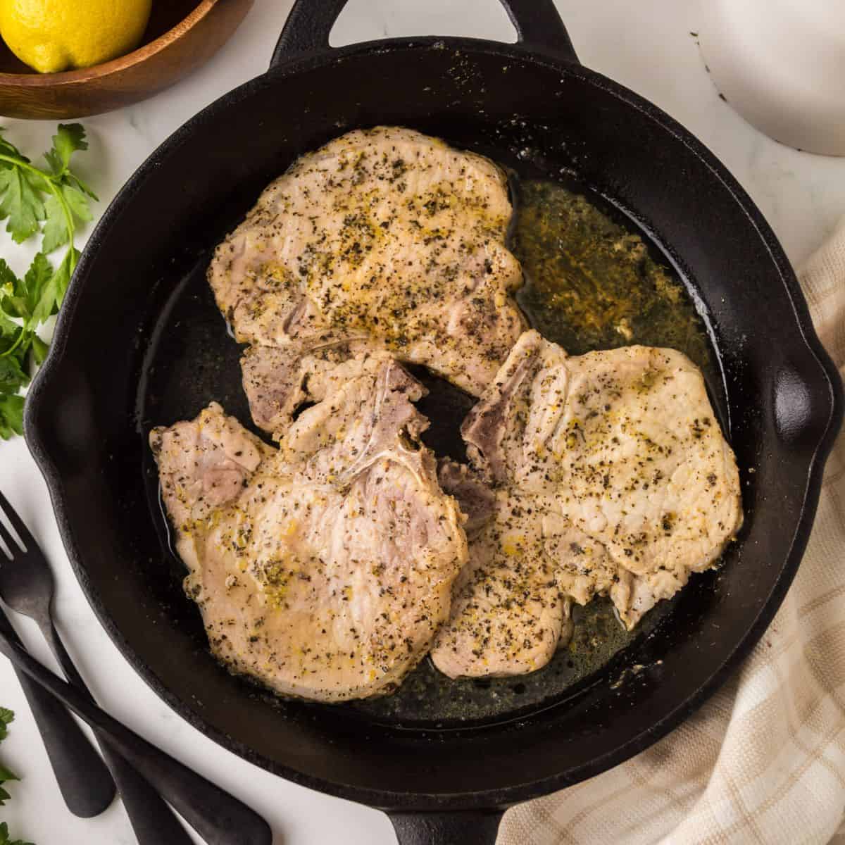 Three seasoned lemon pepper pork chops sizzle in a black cast iron skillet, surrounded by herbs and savory juices. The skillet sits on a white surface with utensils, fresh parsley, and part of a lemon nearby.
